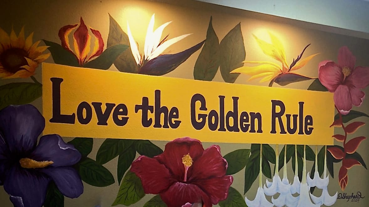 Hand-painted lobby mural reading “Love the Golden Rule,” surrounded by tropical flowers, with pride and transgender flags on the front desk