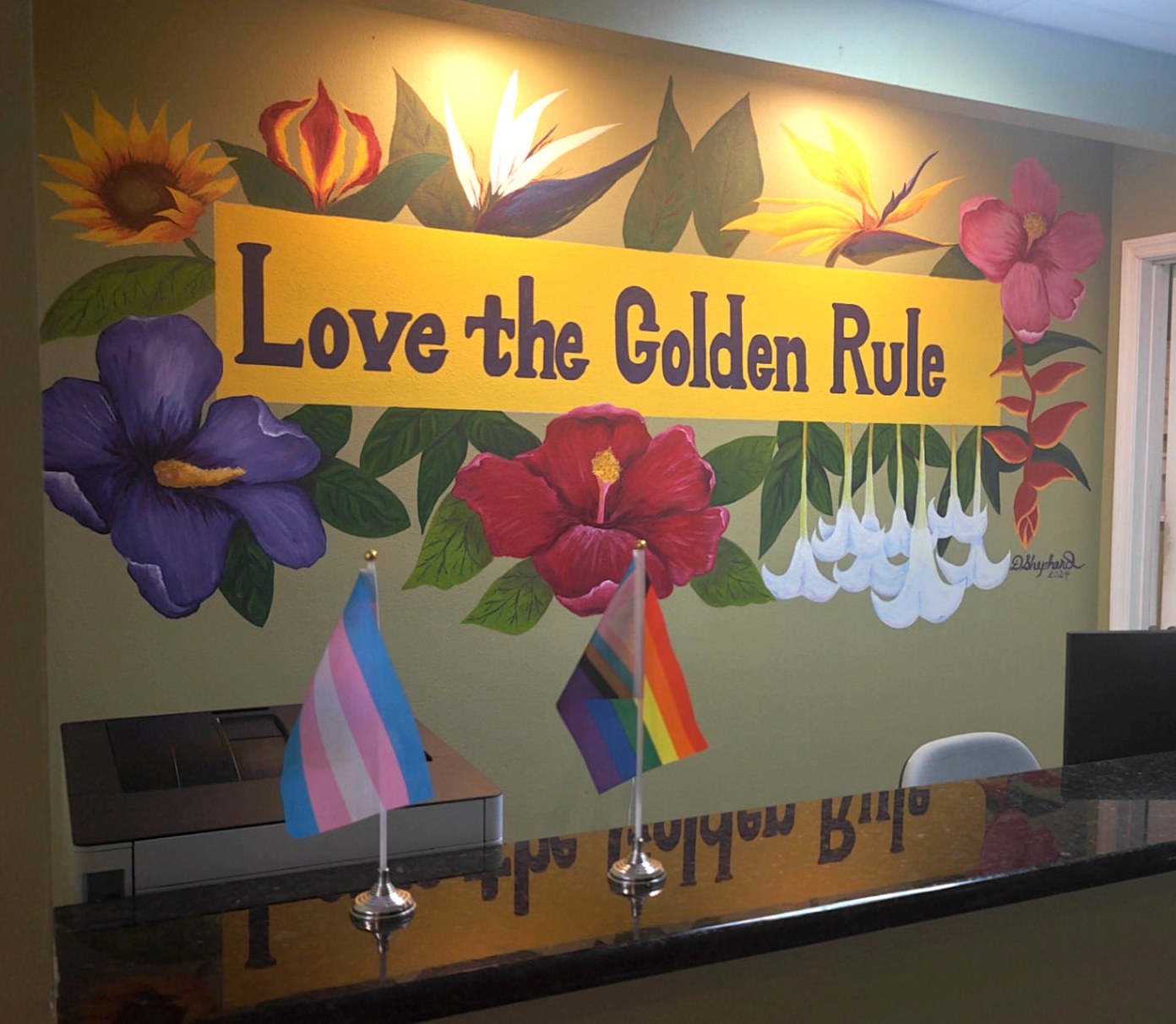 Hand-painted lobby mural reading “Love the Golden Rule,” surrounded by tropical flowers, with pride and transgender flags on the front desk
